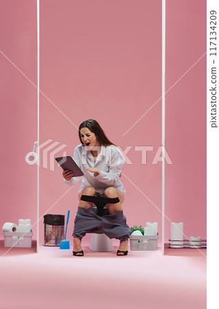 Tense moment. Young woman in white shirt standing in toilet with tablet, watching online game translation with emotional face against pink background. Tense moment. Young woman in white shirt standing in toilet with tablet, watching online game translation with emotional face against pink background. 117134209