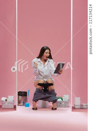 Excited happy young woman, office worker sitting in toilet with tablet and watching soccer game translation against pink background Excited happy young woman, office worker sitting in toilet with tablet and watching soccer game translation against pink background 117134214