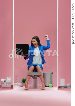 Goal. Emotional businesswoman sitting atop of toilet with laptop and watching live match event, cheering favorite soccer team against pink background Goal. Emotional businesswoman sitting atop of toilet with laptop and watching live match event, cheering favorite soccer team against pink background 117134228