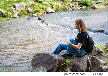 Young energetic girl with backpack having fun on edge of cliff in mountains at sunset against background of clouds. 117134643