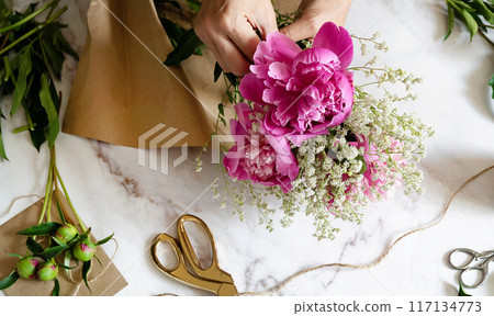 A female florist makes a bouquet of peony flowers wrapped in kraft paper on her desktop. View from above. High quality photos A female florist makes a bouquet of peony flowers wrapped in kraft paper on her desktop. View from above. High quality photos 117134773
