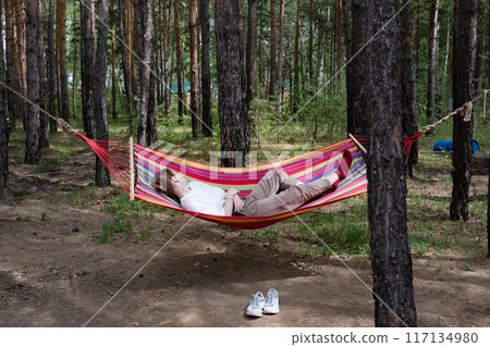 Beautiful young woman relaxing in hammock in forest. Summer scenery, a beautiful morning in the bosom of nature. The girl admires the views and nature. Breathed fresh air. Beautiful morning light. 117134980