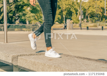 Slender female legs in white sneakers jumping between concrete blocks in a square. Slender female legs in white sneakers jumping between concrete blocks in a square. 117135048