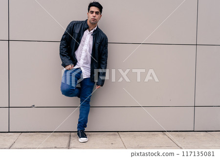 Full body photograph of a young Latino guy leaning against a plain colored wall. Modern urban clothing. 117135081