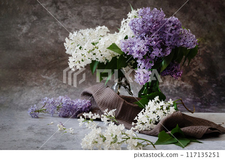 Front view of lilac flowers bouquet in ceramic vase on wooden garden table against blurred outdoor terrace in sunny day Front view of lilac flowers bouquet in ceramic vase on wooden garden table against blurred outdoor terrace in sunny day 117135251