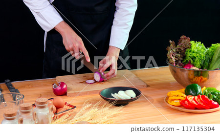 Young Chef chopping cucumber on wooden cutting board in kitchen, surrounded by fresh vegetables, eggs, and spices, preparing food Young Chef chopping cucumber on wooden cutting board in kitchen, surrounded by fresh vegetables, eggs, and spices, preparing food 117135304