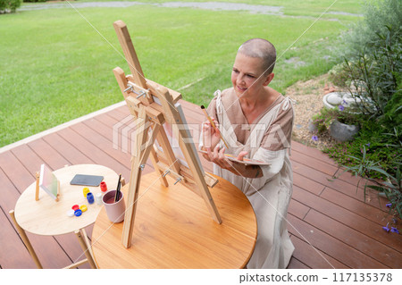 Woman Painting on an Easel Outdoors in a Garden Setting on a Wooden Deck Woman Painting on an Easel Outdoors in a Garden Setting on a Wooden Deck 117135378