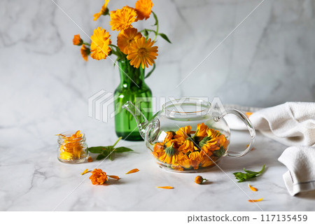Cup of marigold tea and calendula flowers outdoors on green background. Cup of marigold tea and calendula flowers outdoors on green background. 117135459