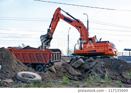 Excavator during earthmoving at open pit on blue sky background. Construction machinery and earth-moving heavy equipment for excavation, loading, lifting and hauling of cargo on job sites Excavator during earthmoving at open pit on blue sky background. Construction machinery and earth-moving heavy equipment for excavation, loading, lifting and hauling of cargo on job sites 117135604