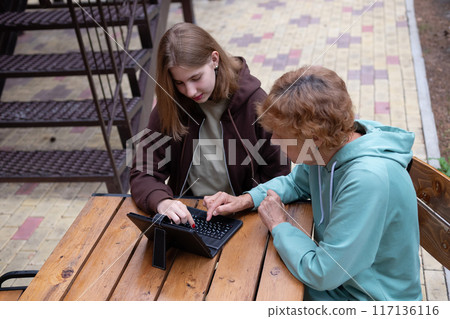 computer awareness of the elderly. a happy teenage girl teaches older 70s grandmothers to use a laptop at home. social media and modern technologies computer awareness of the elderly. a happy teenage girl teaches older 70s grandmothers to use a laptop at home. social media and modern technologies 117136116