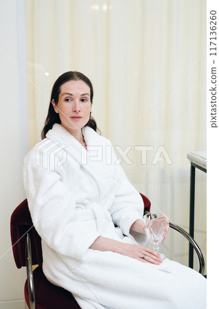 Relaxing Woman in a White Bathrobe Sitting While Holding a Glass in a Bright Indoor Space Relaxing Woman in a White Bathrobe Sitting While Holding a Glass in a Bright Indoor Space 117136260