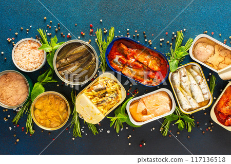 Different open tin cans with canned fish like salmon, mackerel, sprat, sardine, tuna, and herring, among spices and herbs on a blue background Different open tin cans with canned fish like salmon, mackerel, sprat, sardine, tuna, and herring, among spices and herbs on a blue background 117136518