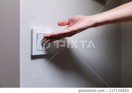 Close-up of an unrecognizable young Caucasian woman's hand turning on a light in a dark room by pressing a switch on a white wall. Close-up Close-up of an unrecognizable young Caucasian woman's hand turning on a light in a dark room by pressing a switch on a white wall. Close-up 117136883