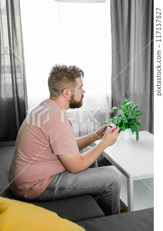 Cheerful young man seated on a sofa playing video games at home. Gaming addiction and entertainment Cheerful young man seated on a sofa playing video games at home. Gaming addiction and entertainment 117137827