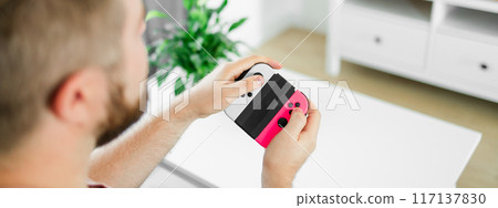 Banner man spending time at home, sitting on a couch in apartment and playing arcade car video games on console copy space. Male using controller to play street racing drift simulator. Banner man spending time at home, sitting on a couch in apartment and playing arcade car video games on console copy space. Male using controller to play street racing drift simulator. 117137830