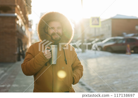 Close-up portrait handsome bearded millennial man in winter clothes and with thermos snow in city copy space. Cold season and hot beverage in winter time 117138697