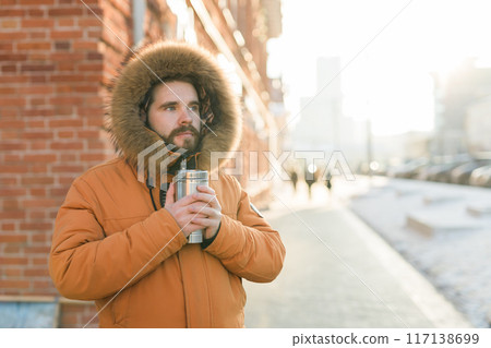 Close-up portrait handsome bearded millennial man in winter clothes and with thermos snow in city copy space. Cold season and hot beverage in winter time Close-up portrait handsome bearded millennial man in winter clothes and with thermos snow in city copy space. Cold season and hot beverage in winter time 117138699