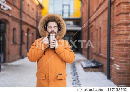 Close-up portrait handsome bearded millennial man in winter clothes and with thermos snow in city. Cold season and hot beverage in winter time Close-up portrait handsome bearded millennial man in winter clothes and with thermos snow in city. Cold season and hot beverage in winter time 117138762