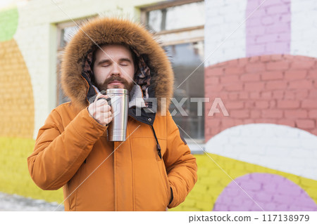 Close-up portrait handsome bearded millennial man in winter clothes and with thermos snow in city copy space. Cold season and hot beverage in winter time 117138979