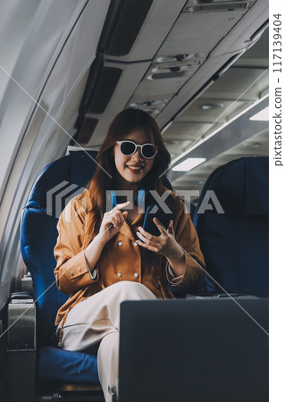 A young Asian woman, seated as an airplane passenger, contemplates dollar bills, reflecting on lucrative opportunities and aspirations towards billionaire status. 117139404