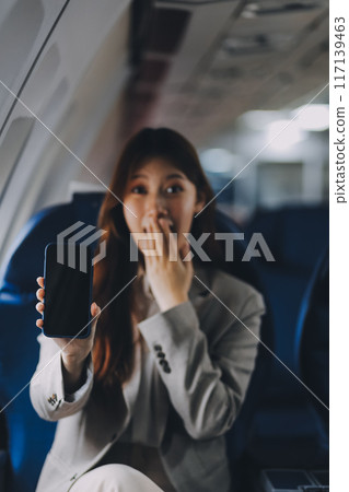 A young Asian woman, seated as an airplane passenger, contemplates dollar bills, reflecting on lucrative opportunities and aspirations towards billionaire status. 117139463