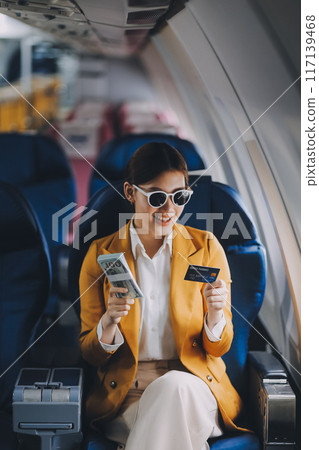 A young Asian woman, seated as an airplane passenger, contemplates dollar bills, reflecting on lucrative opportunities and aspirations towards billionaire status. A young Asian woman, seated as an airplane passenger, contemplates dollar bills, reflecting on lucrative opportunities and aspirations towards billionaire status. 117139468