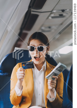 A young Asian woman, seated as an airplane passenger, contemplates dollar bills, reflecting on lucrative opportunities and aspirations towards billionaire status. 117139469