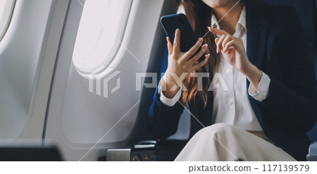 A young Asian female airplane passenger sits by the window during the flight, holding her boarding pass, with her carry-on luggage stored overhead, wearing her seatbelt, and enjoying the journey. A young Asian female airplane passenger sits by the window during the flight, holding her boarding pass, with her carry-on luggage stored overhead, wearing her seatbelt, and enjoying the journey. 117139579