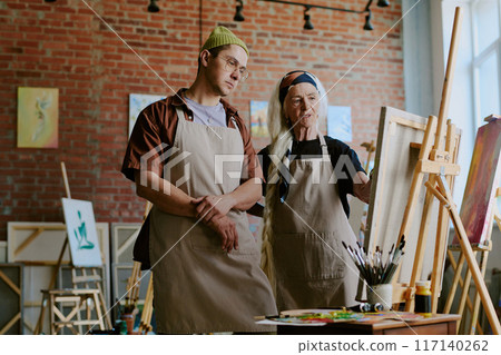 Medium long shot of senior artist helping young man to correct mistake during painting class in studio Medium long shot of senior artist helping young man to correct mistake during painting class in studio 117140262