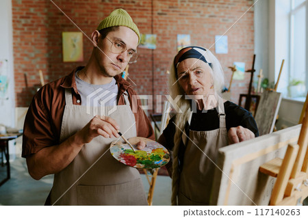 Senior Caucasian woman teaching young man how to paint picture on canvas during individual art class, medium shot 117140263