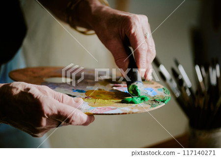 Closeup of hand of unrecognizable artist applying small amount of green oil paint from tube onto palette 117140271
