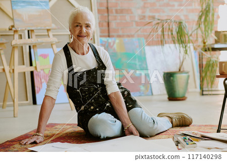 Portrait of creative senior Caucasian woman wearing apron sitting relaxed on floor in modern art studio and smiling at camera 117140298