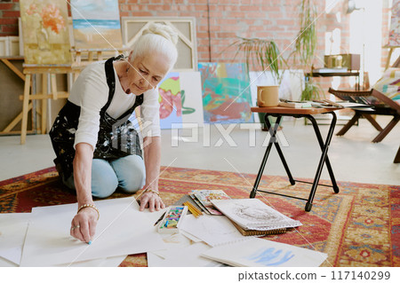 Senior Caucasian female artist sitting on floor in her studio and drawing with soft pastels, copy space Senior Caucasian female artist sitting on floor in her studio and drawing with soft pastels, copy space 117140299