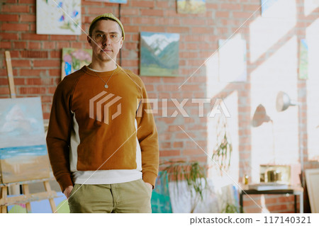 Medium portrait of young Caucasian man wearing casual clothes standing with hands in pockets in his art studio, copy space 117140321