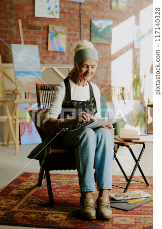 Vertical long shot of elderly Caucasian woman wearing apron sitting on chair in art studio and drawing in sketchbook Vertical long shot of elderly Caucasian woman wearing apron sitting on chair in art studio and drawing in sketchbook 117140328