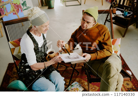 High angle view of young male art teacher working with senior woman looking at portrait sketch in her sketchbook and giving comment High angle view of young male art teacher working with senior woman looking at portrait sketch in her sketchbook and giving comment 117140330