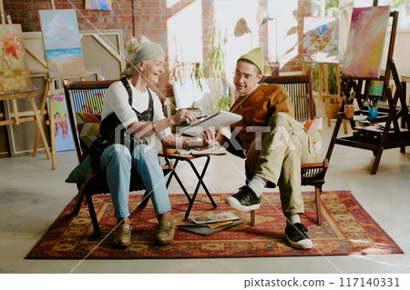 Senior Caucasian female artist sitting on chair in studio showing drawing in sketchbook to young man Senior Caucasian female artist sitting on chair in studio showing drawing in sketchbook to young man 117140331