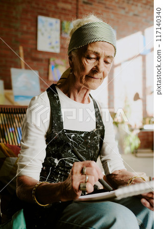 Vertical medium shot of senior Caucasian female artist wearing apron drawing sketch in sketchbook 117140340