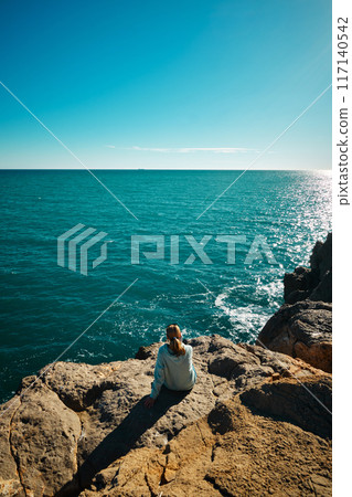 A girl sits on the edge of a cliff and looks at the sea A girl sits on the edge of a cliff and looks at the sea 117140542