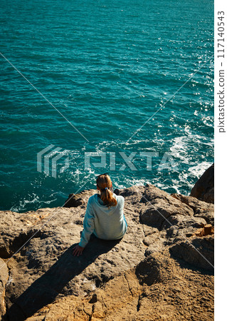 A girl sits on the edge of a cliff and looks at the sea 117140543