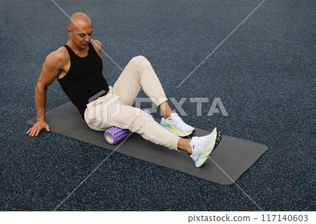 Athlete man exercising and stretching legs on foam roller on sports ground. Outdoor activity. Myofascial Release. 117140603