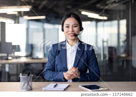 Professional businesswoman in formal attire communicating through a video call in a modern office environment. She is looking at the camera and wearing a headset, conveying professionalism Professional businesswoman in formal attire communicating through a video call in a modern office environment. She is looking at the camera and wearing a headset, conveying professionalism 117140662