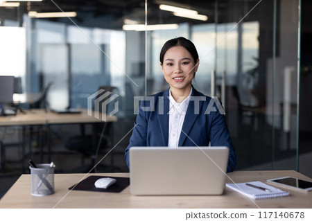 Confident Asian businesswoman working in an office setting, seated at a desk with a laptop, mouse, notepad, and tablet. The professional office environment reflects a productive and modern workspace. 117140678