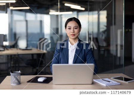 Confident businesswoman in blue suit using laptop at modern office desk. Professional work environment with natural daylight from large windows. Concept of leadership, corporate culture, technology. 117140679