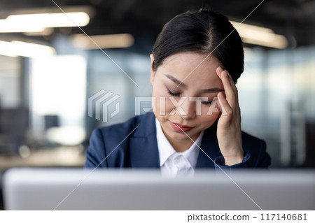 Businesswoman wearing suit feeling stressed and overwhelmed at work. Office worker experiencing exhaustion and frustration in modern office environment. Concept of stress, workplace challenges 117140681