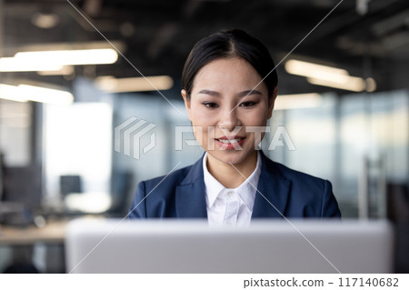 Smiling businesswoman working on laptop in modern office environment. Professional woman in formal attire focused on computer screen, representing productivity and corporate culture in workplace. Smiling businesswoman working on laptop in modern office environment. Professional woman in formal attire focused on computer screen, representing productivity and corporate culture in workplace. 117140682