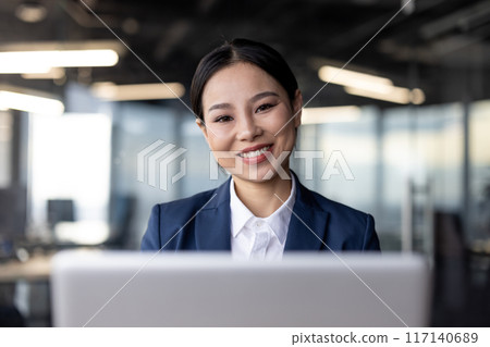Asian businesswoman in formal attire working on a laptop in a modern office setting. She is smiling confidently, embodying professionalism and positivity. 117140689