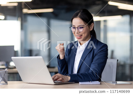 Asian businesswoman working in a modern office environment, using a laptop and celebrating success. Professional attire and enthusiasm show her dedication and achievement in a busy corporate setting. Asian businesswoman working in a modern office environment, using a laptop and celebrating success. Professional attire and enthusiasm show her dedication and achievement in a busy corporate setting. 117140699