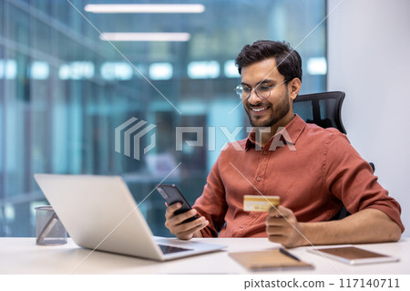 Young professional in a modern office using a phone and credit card for an online transaction. Displaying happiness and success in a business environment. Young professional in a modern office using a phone and credit card for an online transaction. Displaying happiness and success in a business environment. 117140711