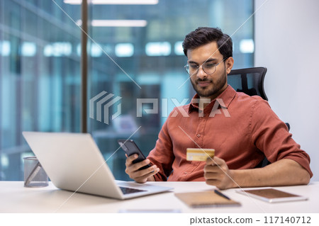 Businessman in orange shirt working in office using smartphone and credit card. Professional setting with laptop, tablet, and office supplies. Modern office environment, online shopping Businessman in orange shirt working in office using smartphone and credit card. Professional setting with laptop, tablet, and office supplies. Modern office environment, online shopping 117140712